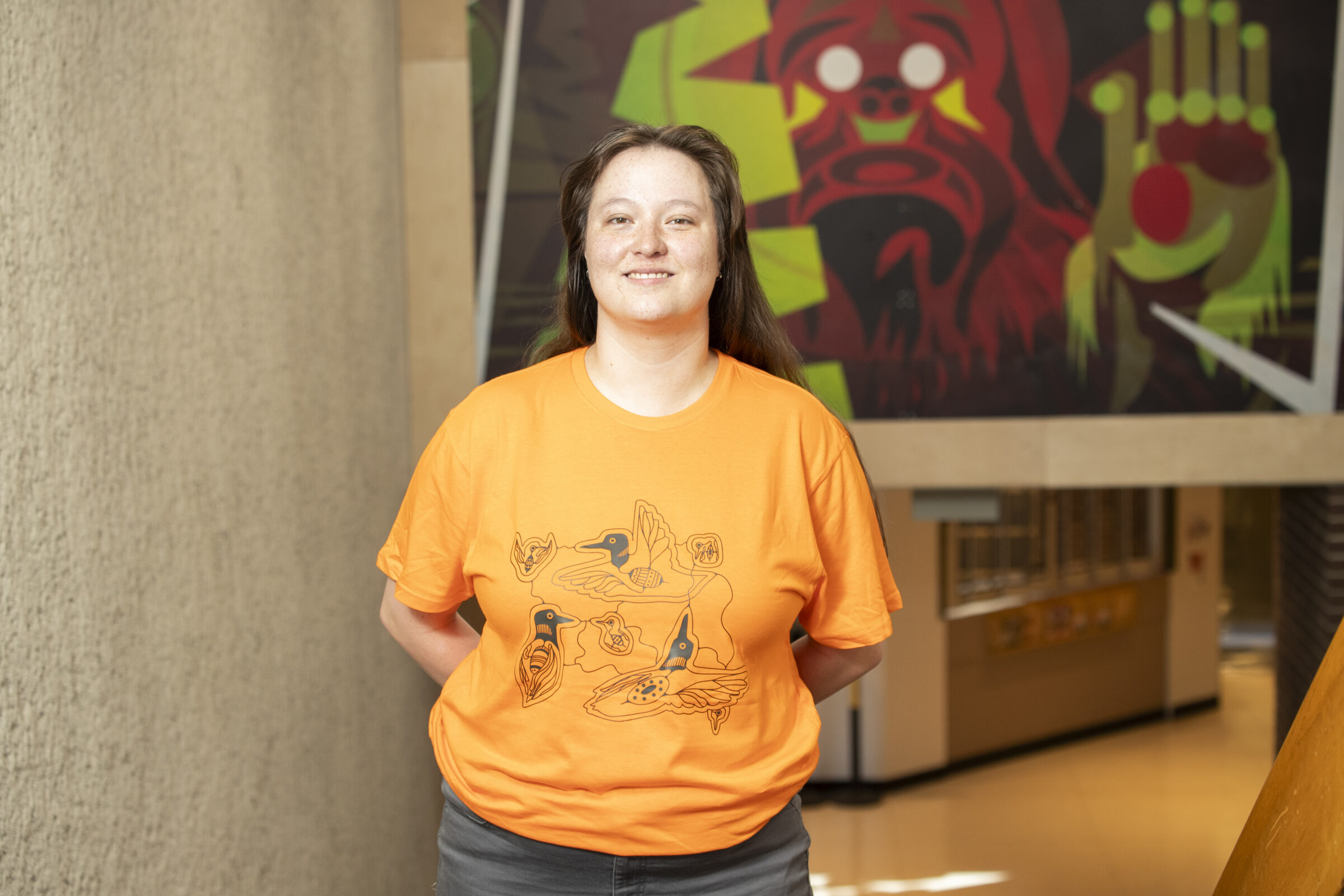 Digital Media Design graduate Erin Ringland stands proudly in front of an Indigenous mural at RRC Polytech's Notre Dame Campus. She is wearing an orange shirt featuring a black loon design for Orange Shirt Day 2024. Erin designed the shirt.