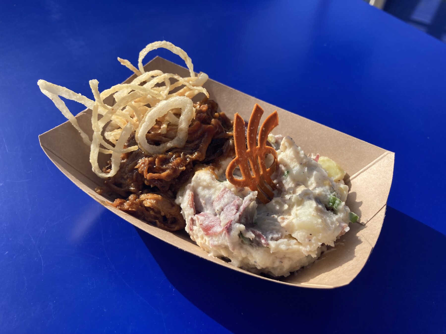 A container filled with pulled pork, crispy onions, and potato salad, topped with an RRC Polytech cracker.