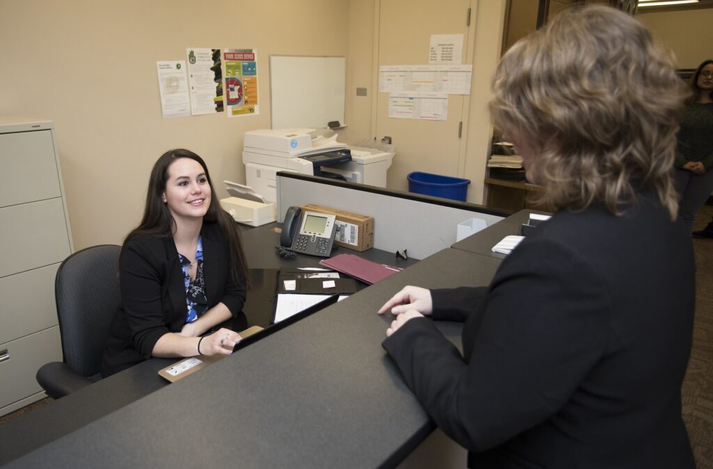 Female administrative assistant speaks to co-worker from behind desk.