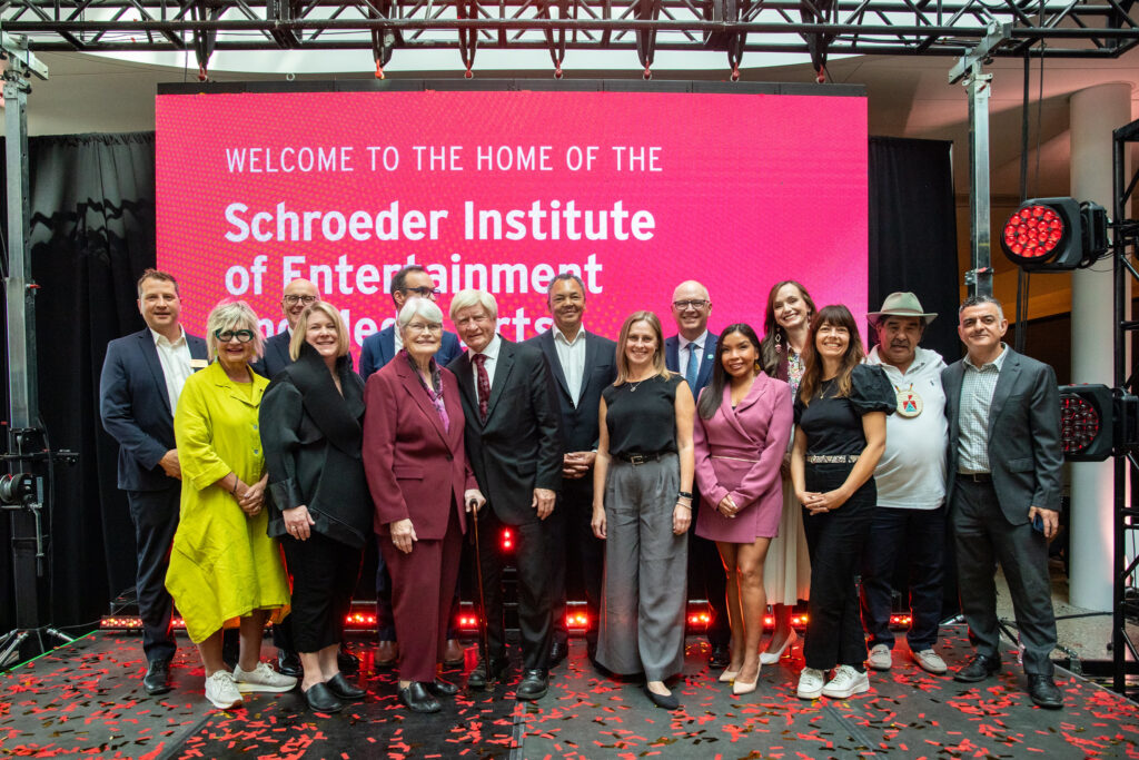 Group shot of Walter and Maria Schroeder, RRC Polytech President Fred Meier, and other College and industry stakeholders, in front of a digital screen announcing RRC Polytech as the home of the Schroeder Institute of Entertainment and Media Arts.