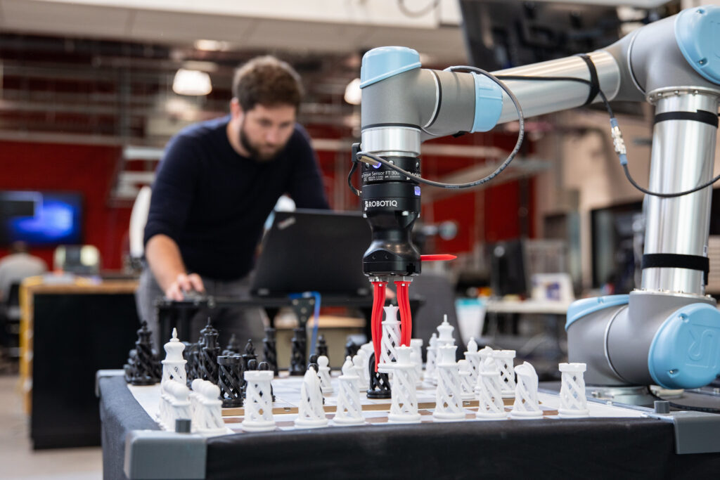 Male RRC Polytech researcher works controls to move a robotic arm clasping a chess piece.