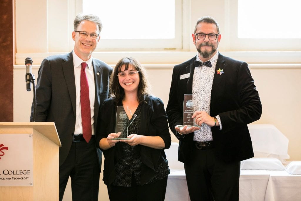 Nora Sobel and Bradley West hold up their glass BRAVO awards in front of a podium with RRC Polytech President Paul Vogt.