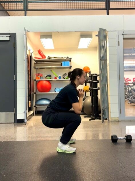  A person in all black is in the fitness centre performing a squat while holding a dumbbell at their chest.