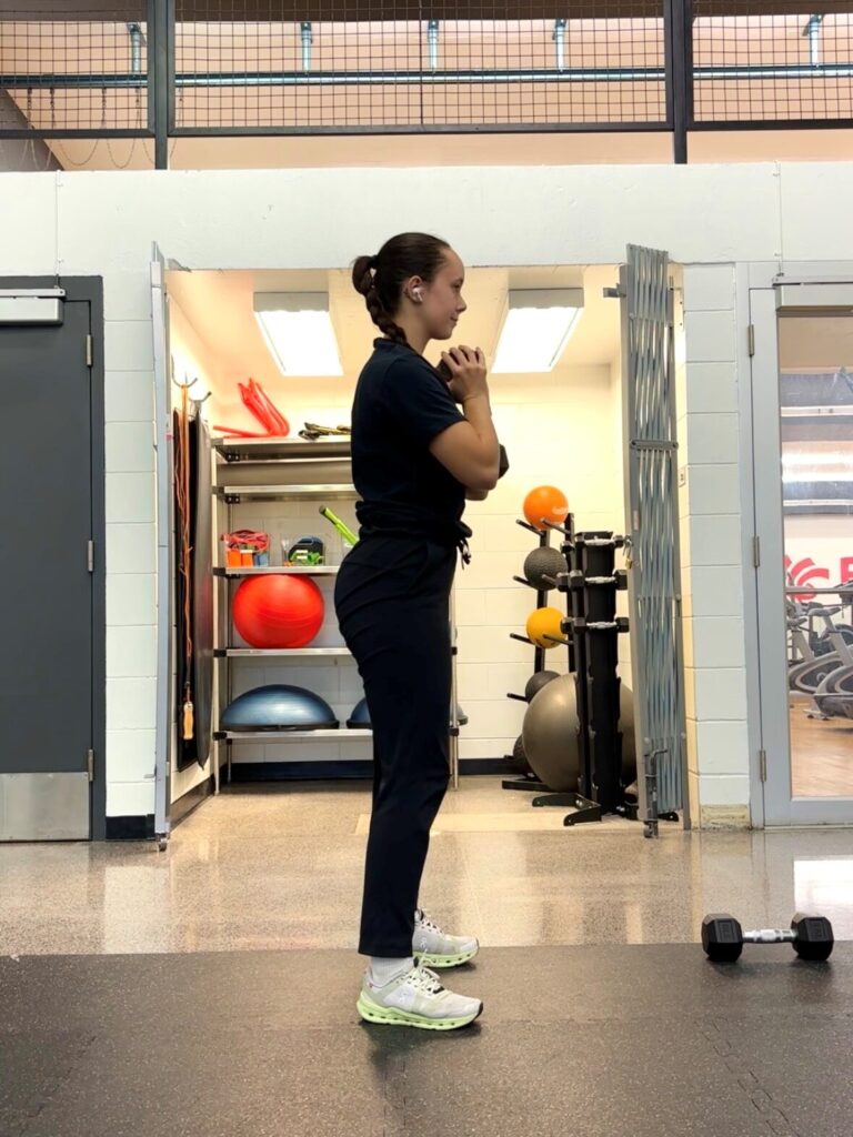 A person in all black is standing in a fitness centre holding a dumbbell at their chest with both hands.