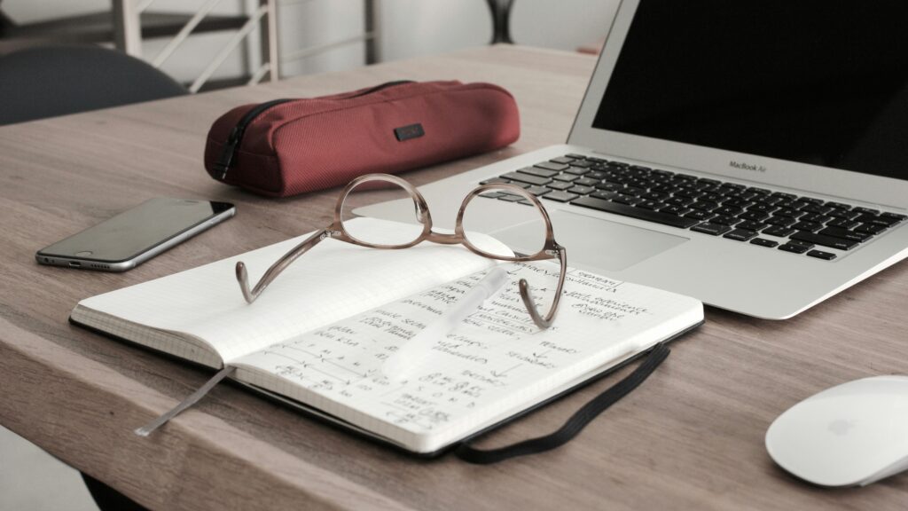 Open computer and notebook with glasses and calculator on a desk.