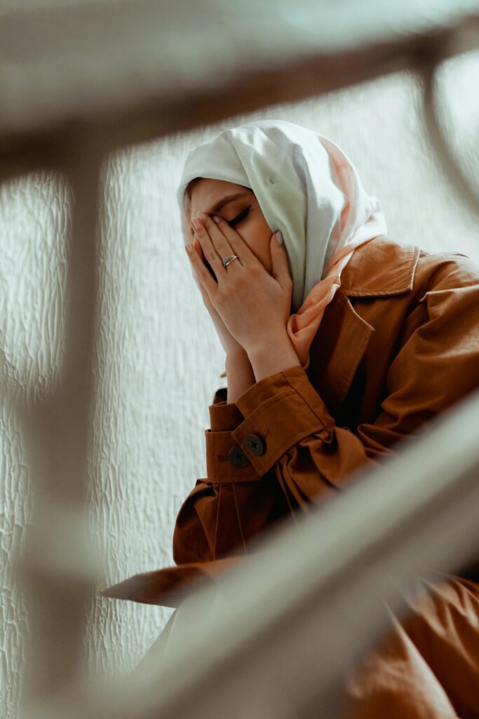 A crying woman with her face in her hands wearing a hijab and coat.