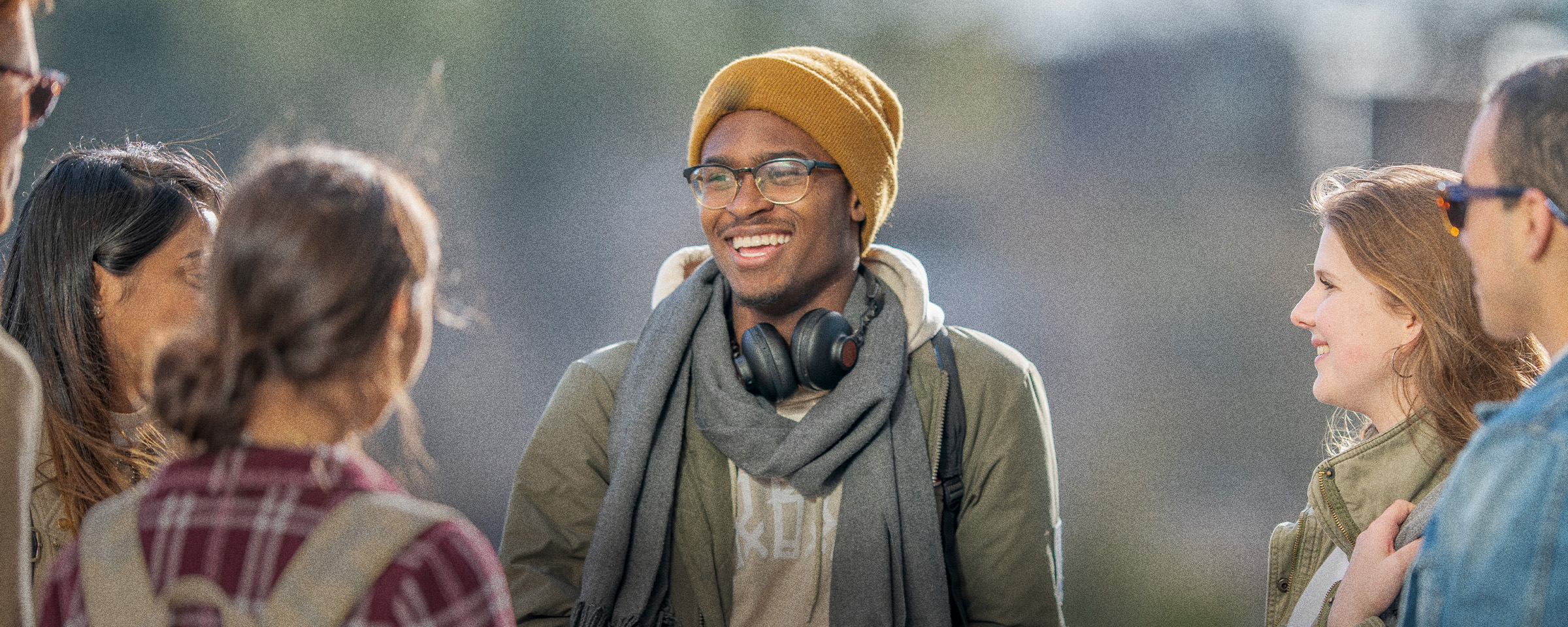 Male student, wearing tuque and headphones, talking to classmates outside.