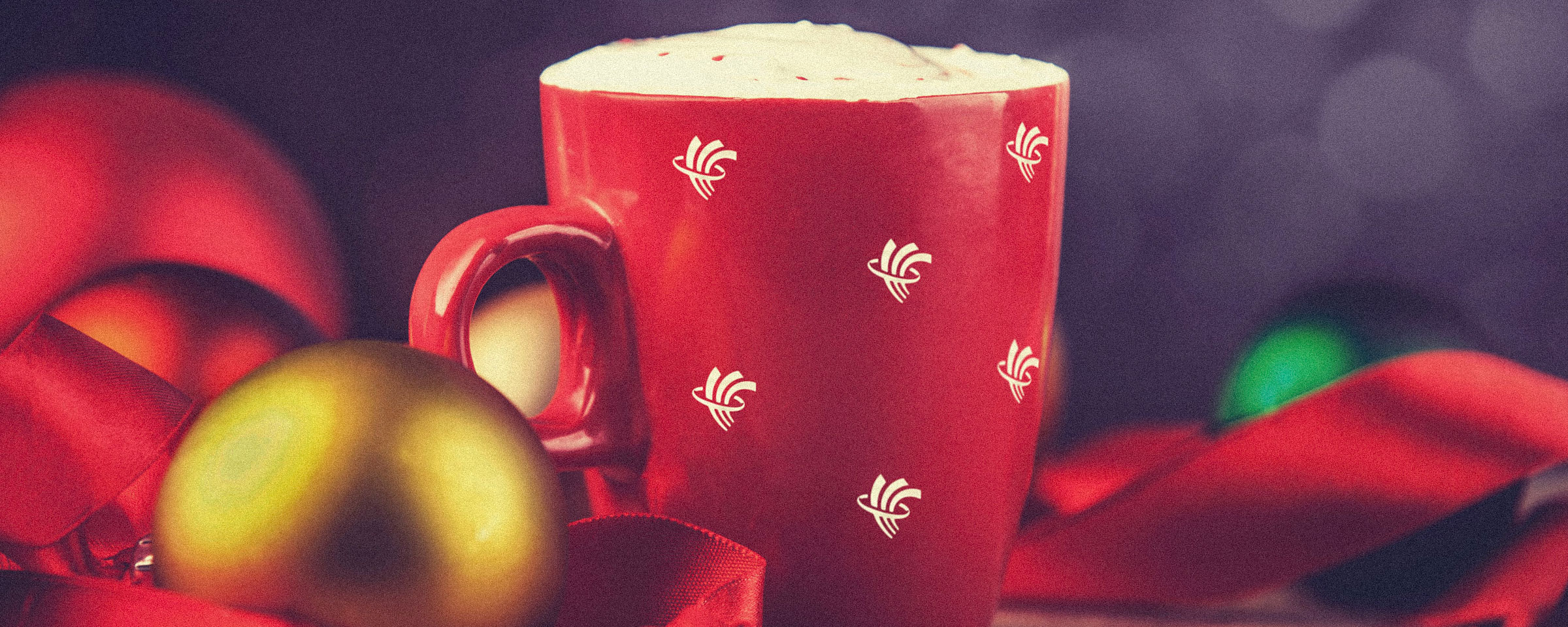 Red mug with elements of RRC logo, filled with hot cocoa and surrounded by holiday decorations and ribbon.